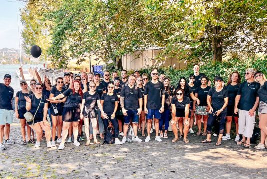 Gruppenfoto von unserem Team in Shorts und schwarzem Coopers Shirt vor dem Zürich See, viele lachende Gesichter