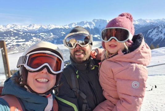 Glückliche Familie beim Skifahren in den österreichischen Alpen. Ein Mann, eine Frau und ihr kleines Kind tragen Skibrillen und warme Winterkleidung, während sie vor einer beeindruckenden Schneelandschaft für ein Foto lächeln.