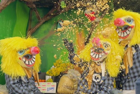 Three carnival participants with large, colorful masks, yellow hair and checkered costumes throw confetti into the air, typical of the Basel carnival.