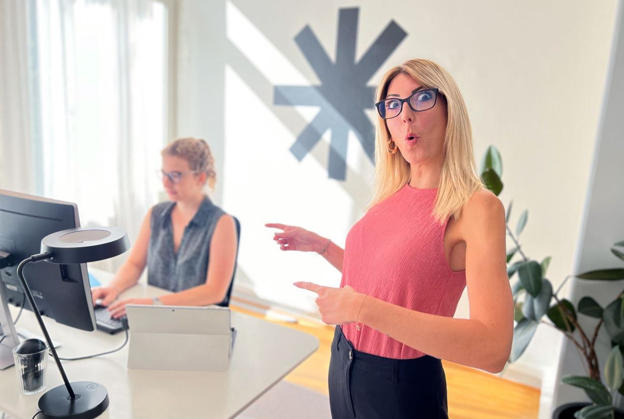 Women pointing to another women who sits and works at the computer at the desk