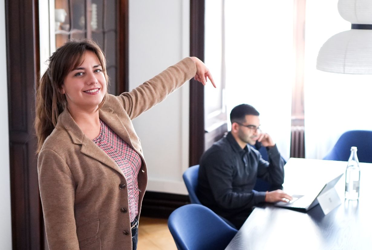 Two people, one standing and pointing at the person sitting at the table.